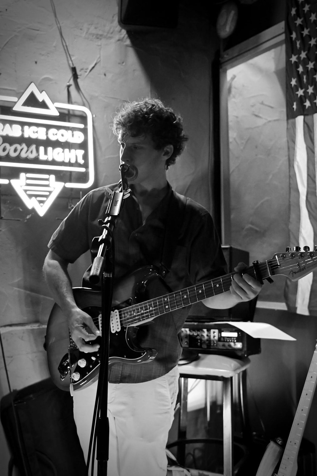 Black and white photo of Austin Finnegan singing and playing guitar by a neon sign at Sierra Madre Saloon