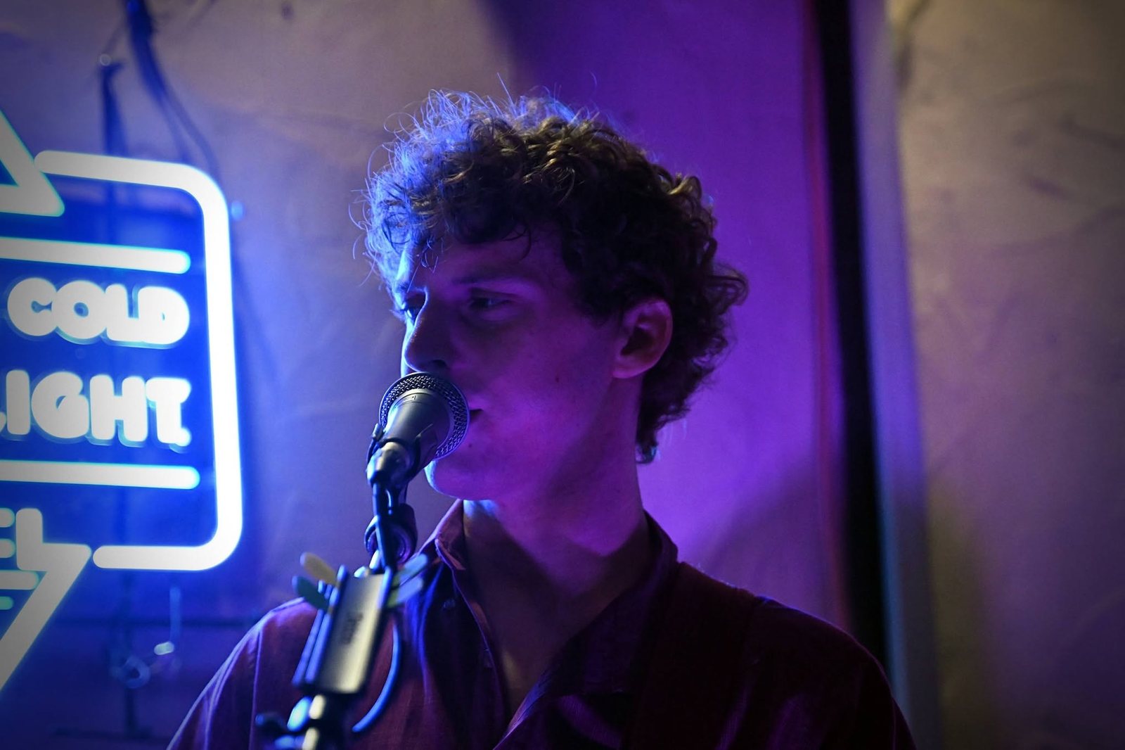Close-up of Austin Finnegan singing into the mic under purple light beside a neon sign at Sierra Madre Saloon